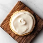 Shortbread cookie icing - the image shows a round cookie on a wooden cutting board. the cookie appears to be a shortbread cookie with a golden brown crust. on top of the cookie, there is a generous layer of white frosting. the frosting is spread evenly across the cookie and has a smooth texture. the cutting board is placed on a white marble surface.