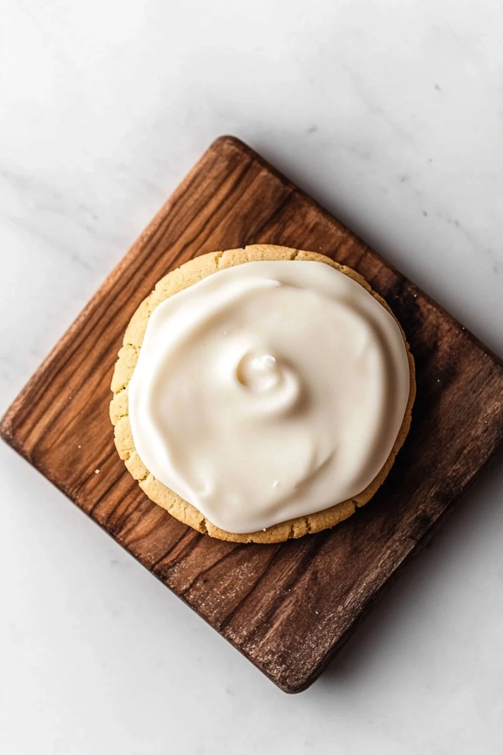 Shortbread cookie icing - the image shows a round cookie on a wooden cutting board. the cookie appears to be a shortbread cookie with a golden brown crust. on top of the cookie, there is a generous layer of white frosting. the frosting is spread evenly across the cookie and has a smooth texture. the cutting board is placed on a white marble surface.