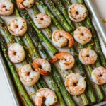 Shrimp sheet pan with asparagus - the image shows a baking tray filled with cooked shrimp and asparagus. the shrimp are arranged in a single layer on the tray, with some of them overlapping each other. the tray is lined with parchment paper and there are a few lemon wedges scattered around the tray. the background is a white wooden table.