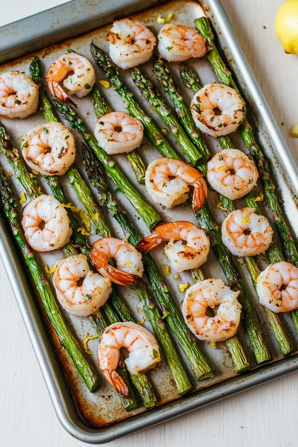 Shrimp sheet pan with asparagus - the image shows a baking tray filled with cooked shrimp and asparagus. the shrimp are arranged in a single layer on the tray, with some of them overlapping each other. the tray is lined with parchment paper and there are a few lemon wedges scattered around the tray. the background is a white wooden table.