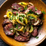 Skillet beef liver - the image is a close-up of a plate of food. the plate is orange in color and has a speckled texture. on top of the plate, there are several pieces of cooked steak, which appear to be medium-rare and have a pinkish-red color. the steak is garnished with sliced yellow bell peppers, onions, and chopped herbs. the dish is served on a black textured surface. the overall appearance of the dish is savory and appetizing.