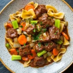 Slow cooker beef with noodles - the image is a close-up of a bowl of beef noodle soup. the bowl is made of ceramic and has a light brown color with a speckled pattern. the soup is made up of beef, carrots, celery, and noodles. the beef is cooked in a rich, dark brown sauce and is garnished with chopped herbs. the noodles are long and thin, and the vegetables are bright orange and green. the dish is served on a dark grey background.