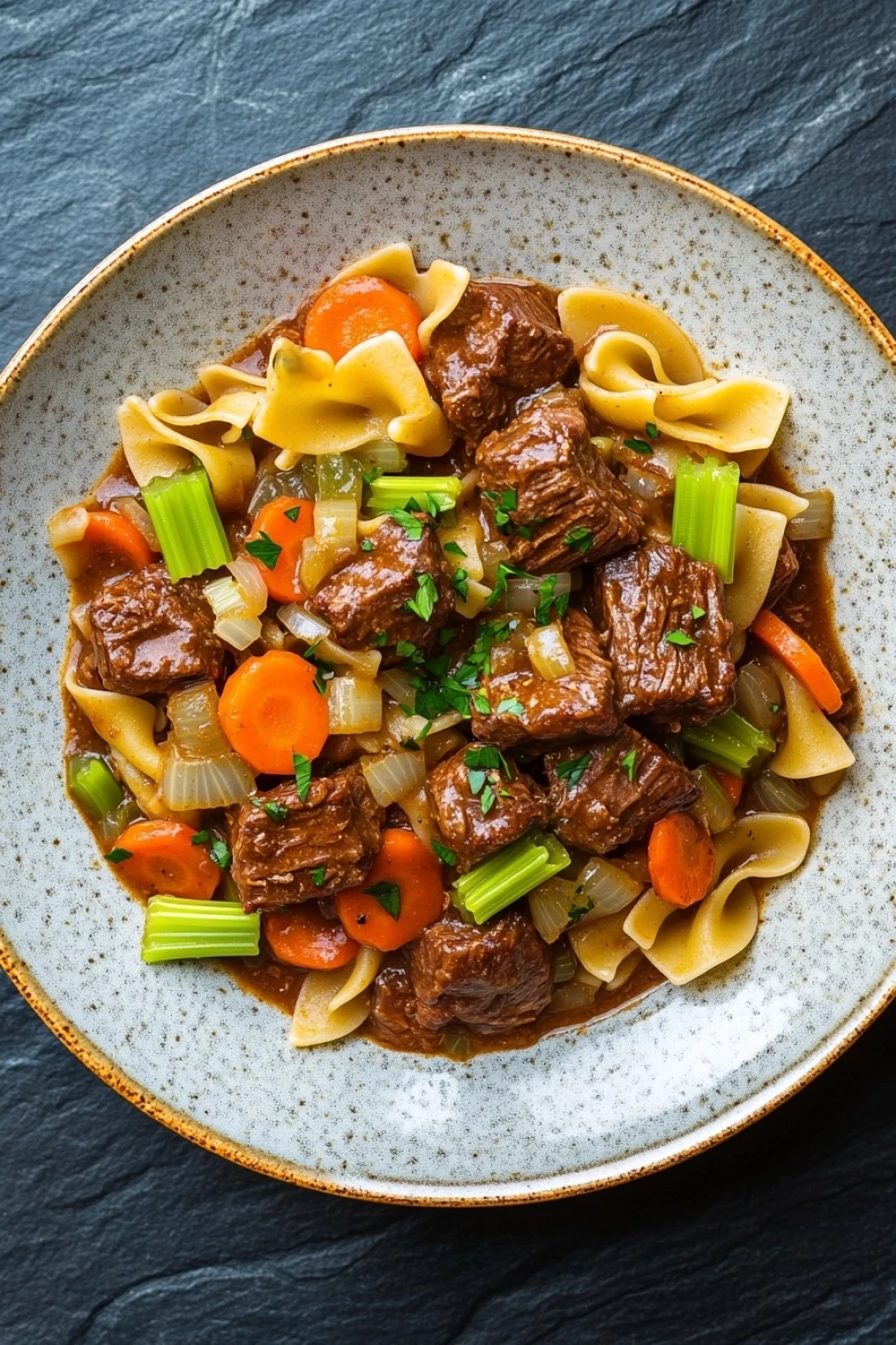 Slow cooker beef with noodles - the image is a close-up of a bowl of beef noodle soup. the bowl is made of ceramic and has a light brown color with a speckled pattern. the soup is made up of beef, carrots, celery, and noodles. the beef is cooked in a rich, dark brown sauce and is garnished with chopped herbs. the noodles are long and thin, and the vegetables are bright orange and green. the dish is served on a dark grey background.