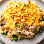 Tuna casserole slow cooker - the image is a close-up of a plate of macaroni and cheese. the plate is round and has a light brown color with a speckled pattern. on top of the macaronis, there is a layer of melted cheese with crumbled breadcrumbs scattered on top. the cheese is a bright yellow color and appears to be melted and bubbly. there are also small pieces of chicken and peas scattered throughout the dish. the dish is garnished with chopped parsley. the background is a white marble countertop.