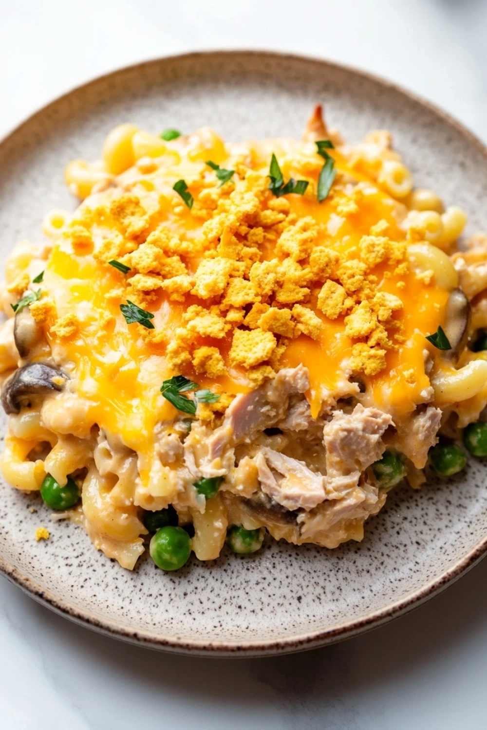 Tuna casserole slow cooker - the image is a close-up of a plate of macaroni and cheese. the plate is round and has a light brown color with a speckled pattern. on top of the macaronis, there is a layer of melted cheese with crumbled breadcrumbs scattered on top. the cheese is a bright yellow color and appears to be melted and bubbly. there are also small pieces of chicken and peas scattered throughout the dish. the dish is garnished with chopped parsley. the background is a white marble countertop.
