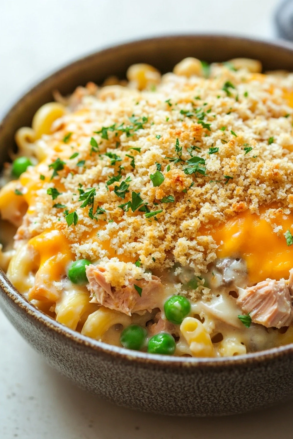 Tuna casserole without soup - the image is a close-up of a bowl of macaroni and cheese. the bowl is made of ceramic and has a dark brown color. the top of the dish is covered in a layer of breadcrumbs, which are sprinkled with chopped parsley. the cheese is melted and bubbly, and there are chunks of salmon and peas scattered throughout the dish. the dish appears to be freshly cooked and ready to eat.