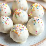Vanilla cake balls - the image shows a plate of white chocolate truffles with colorful sprinkles on top. the plate is round and has a light blue rim with a speckled pattern. the truffels are round and have a smooth texture. they are arranged in a scattered manner on the plate, with some overlapping each other. the sprinkles are in various colors, including red, orange, yellow, green, blue, and purple. the background is blurred, but it appears to be a white wooden table.