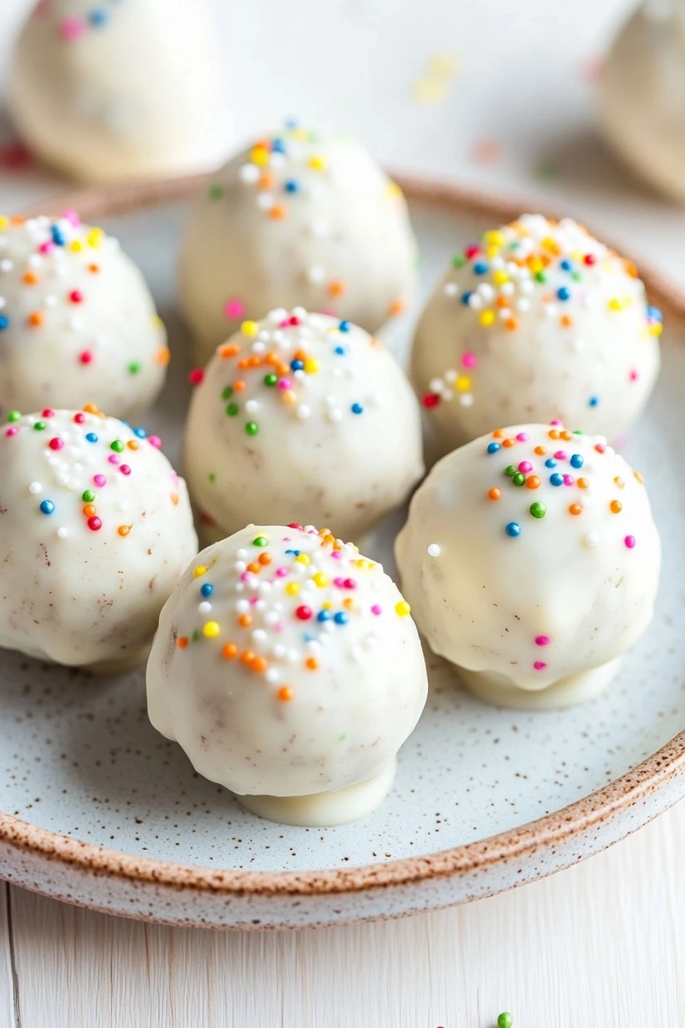 Vanilla cake balls - the image shows a plate of white chocolate truffles with colorful sprinkles on top. the plate is round and has a light blue rim with a speckled pattern. the truffels are round and have a smooth texture. they are arranged in a scattered manner on the plate, with some overlapping each other. the sprinkles are in various colors, including red, orange, yellow, green, blue, and purple. the background is blurred, but it appears to be a white wooden table.