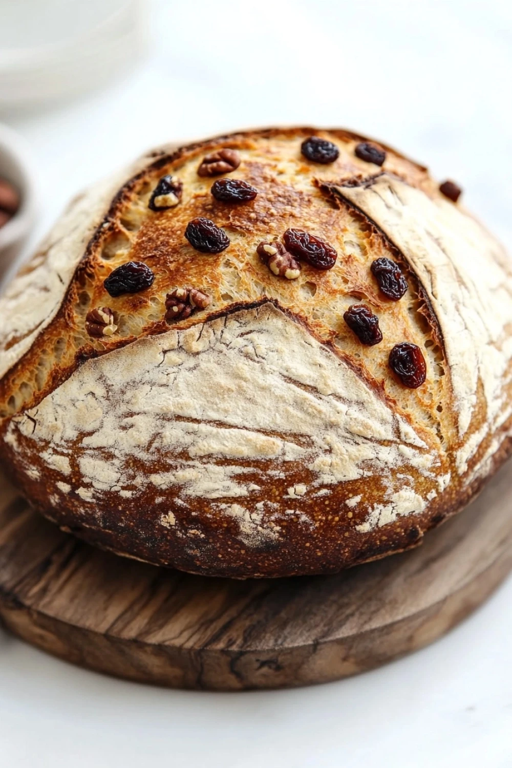 Yeast bread with inclusions - the image shows a round loaf of bread on a wooden cutting board. the bread appears to be freshly baked and has a golden brown crust on top. it is topped with sliced raisins and pecans, giving it a crunchy texture. the cutting board is placed on a white marble countertop, and there is a small bowl of nuts in the background. the overall aesthetic of the image is rustic and homey.