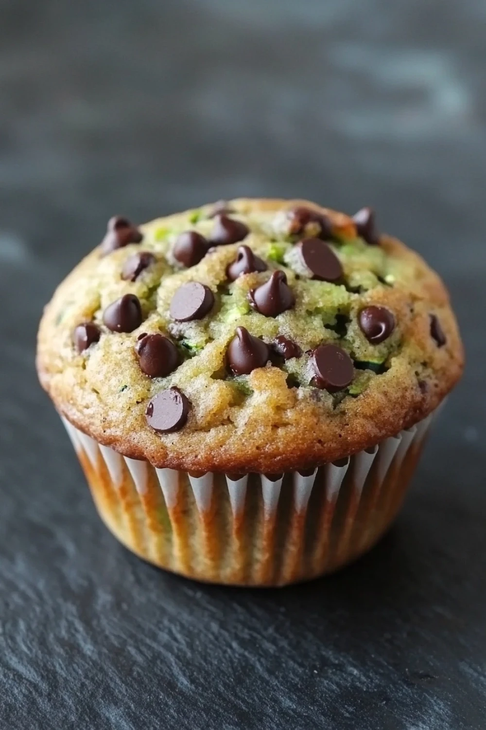 Zucchini banana chocolate chip muffin - the image is of a freshly baked muffin on a black slate surface. the muffin is golden brown in color and has a crumbly texture. it is topped with chocolate chips scattered across the top, giving it a crunchy appearance. the chocolate chips are dark brown and appear to be freshly baked. the background is blurred, making the muffin the focal point of the image.
