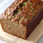 Zucchini bread no sugar - the image shows a freshly baked loaf of zucchini bread on a wooden cutting board. the bread is golden brown in color and has a crumbly texture. it appears to be moist and fluffy, with visible zucchinis and herbs scattered throughout. the cutting board is placed on a white wooden table with a rustic background.