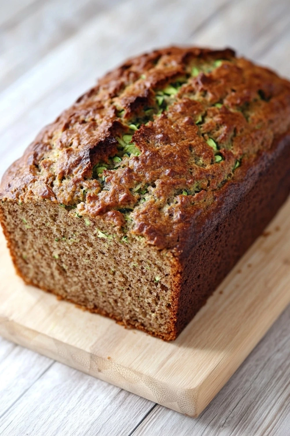 Zucchini bread no sugar - the image shows a freshly baked loaf of zucchini bread on a wooden cutting board. the bread is golden brown in color and has a crumbly texture. it appears to be moist and fluffy, with visible zucchinis and herbs scattered throughout. the cutting board is placed on a white wooden table with a rustic background.